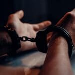 A close-up of human hands handcuffed on a table with a dark background, depicting law enforcement themes.